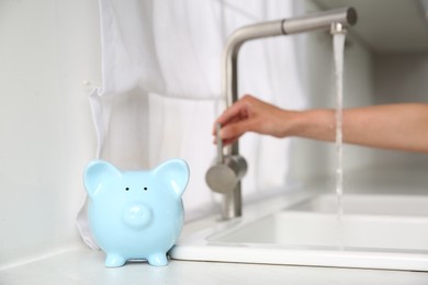 Photo of Water saving concept. Woman checking water flow in kitchen, focus on piggy bank