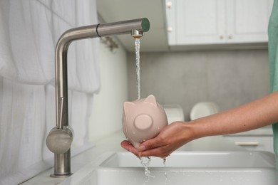 Photo of Water saving concept. Woman with piggy bank under tap water in kitchen