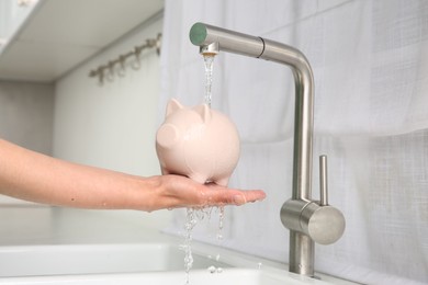 Photo of Water saving concept. Woman with piggy bank under tap water in kitchen