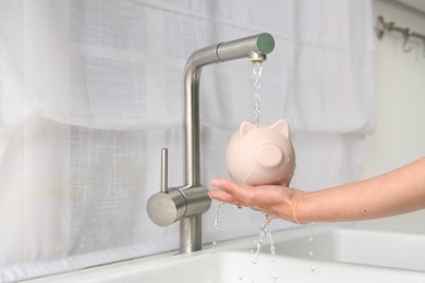 Photo of Water saving concept. Woman with piggy bank under tap water in kitchen