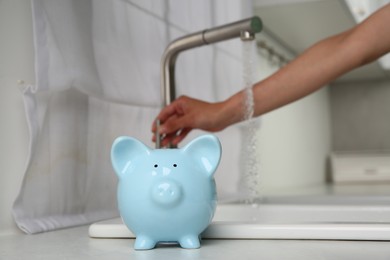 Photo of Water saving concept. Woman checking water flow in kitchen, focus on piggy bank