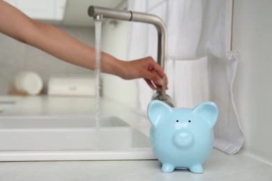 Photo of Water saving concept. Woman checking water flow in kitchen, focus on piggy bank