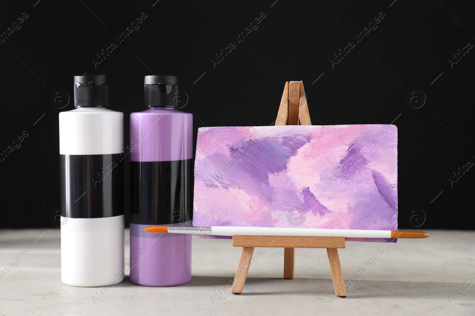 Photo of Acrylic paints of different colors in bottles, brush and small easel with abstract picture on light table