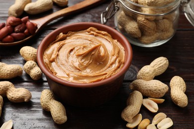 Photo of Tasty peanut butter in bowl, groundnuts, jar and spoon on wooden table, closeup