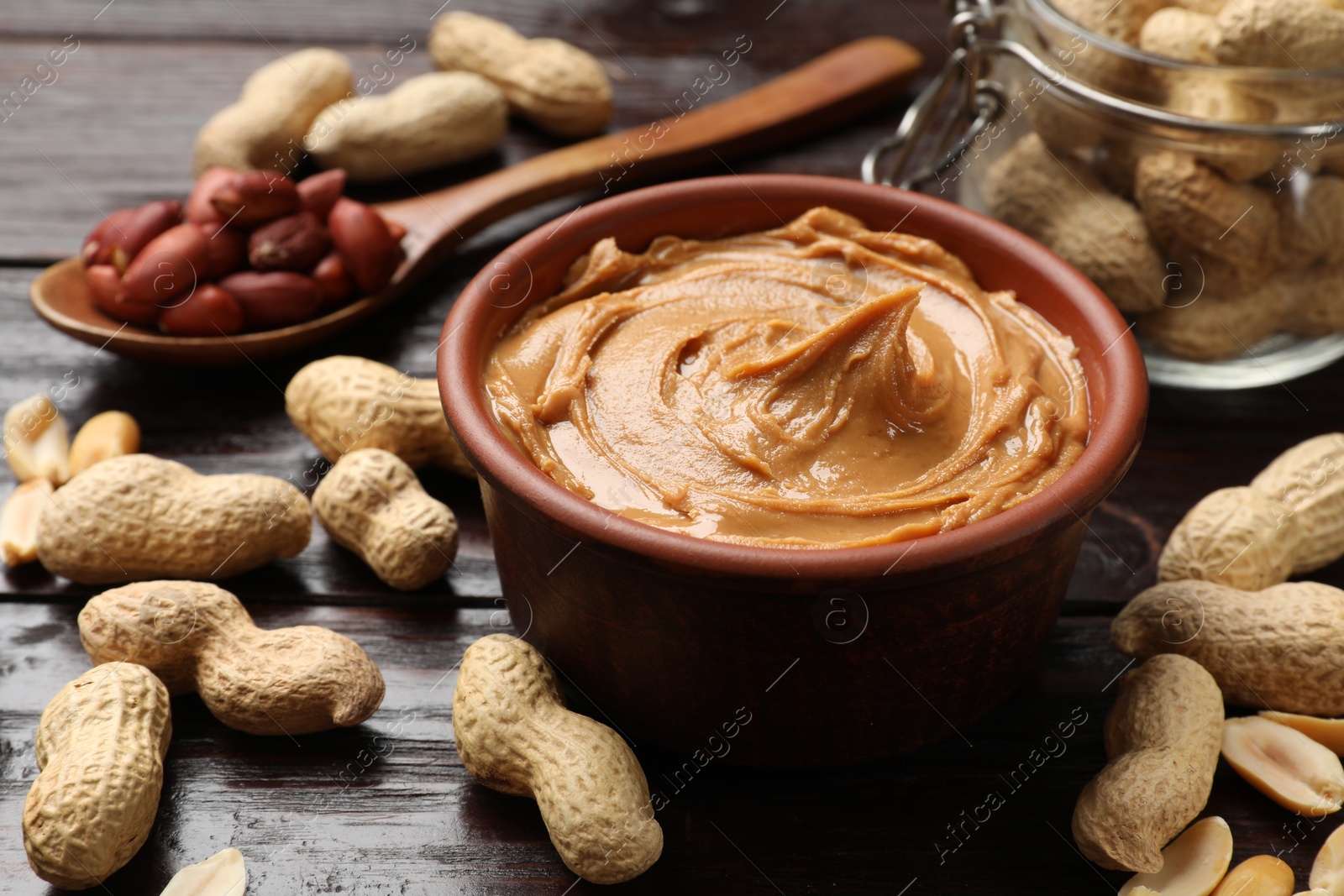 Photo of Tasty peanut butter in bowl, groundnuts, jar and spoon on wooden table, closeup