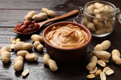 Photo of Tasty peanut butter in bowl, groundnuts, jar and spoon on wooden table, closeup