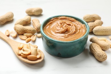 Photo of Tasty peanut butter in bowl and spoon with groundnuts on white table, closeup