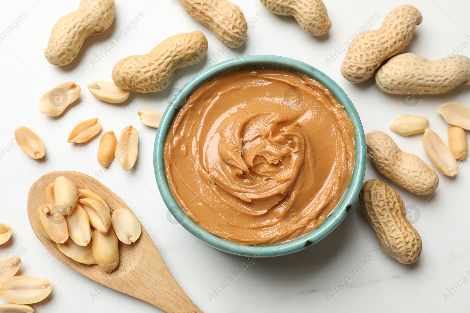 Photo of Tasty peanut butter in bowl and spoon with groundnuts on white table, flat lay