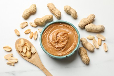 Photo of Tasty peanut butter in bowl and spoon with groundnuts on white table, flat lay