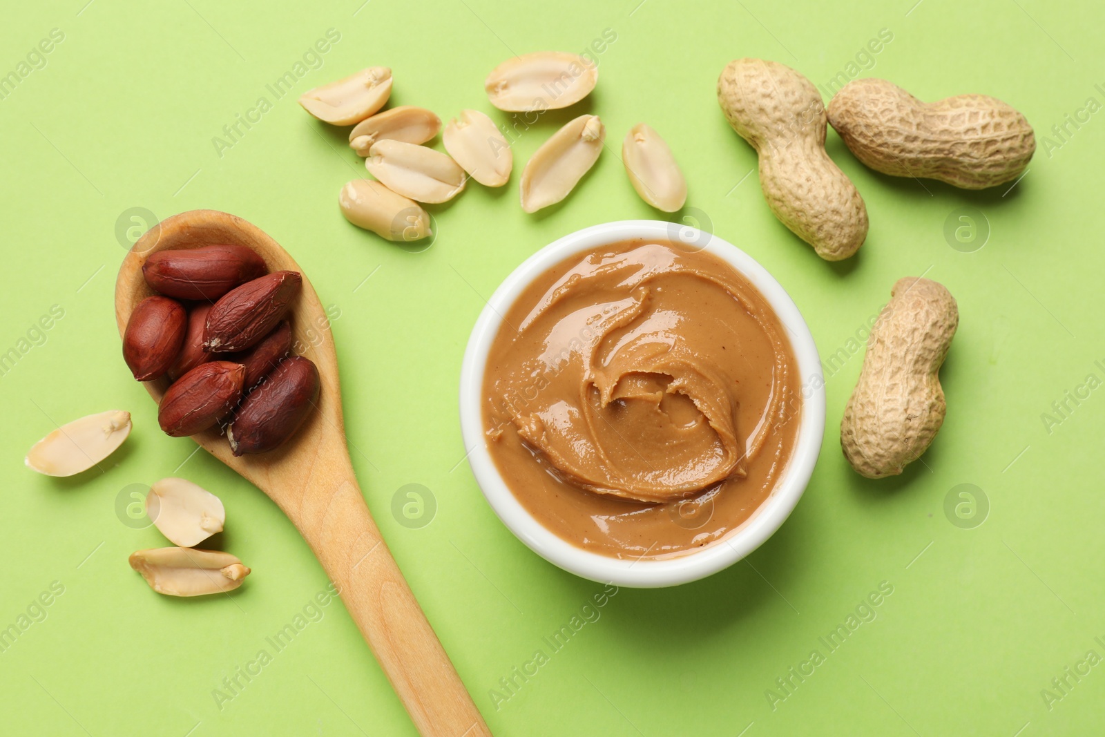 Photo of Tasty peanut butter in bowl and spoon with groundnuts on green table, flat lay