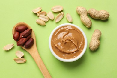 Photo of Tasty peanut butter in bowl and spoon with groundnuts on green table, flat lay