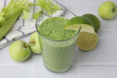 Photo of Tasty green smoothie in glass with lime and apples on white tiled table, closeup