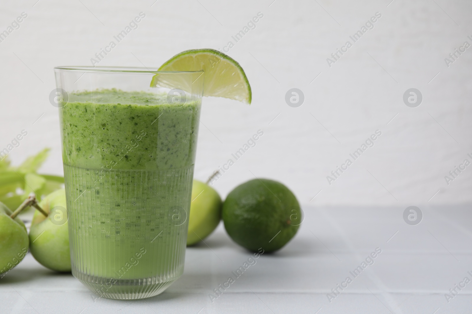 Photo of Tasty green smoothie in glass with lime and apples on white tiled table, closeup. Space for text