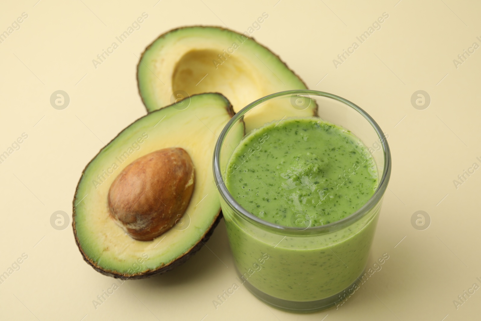 Photo of Tasty green smoothie in glass and halves of avocado on beige background, closeup