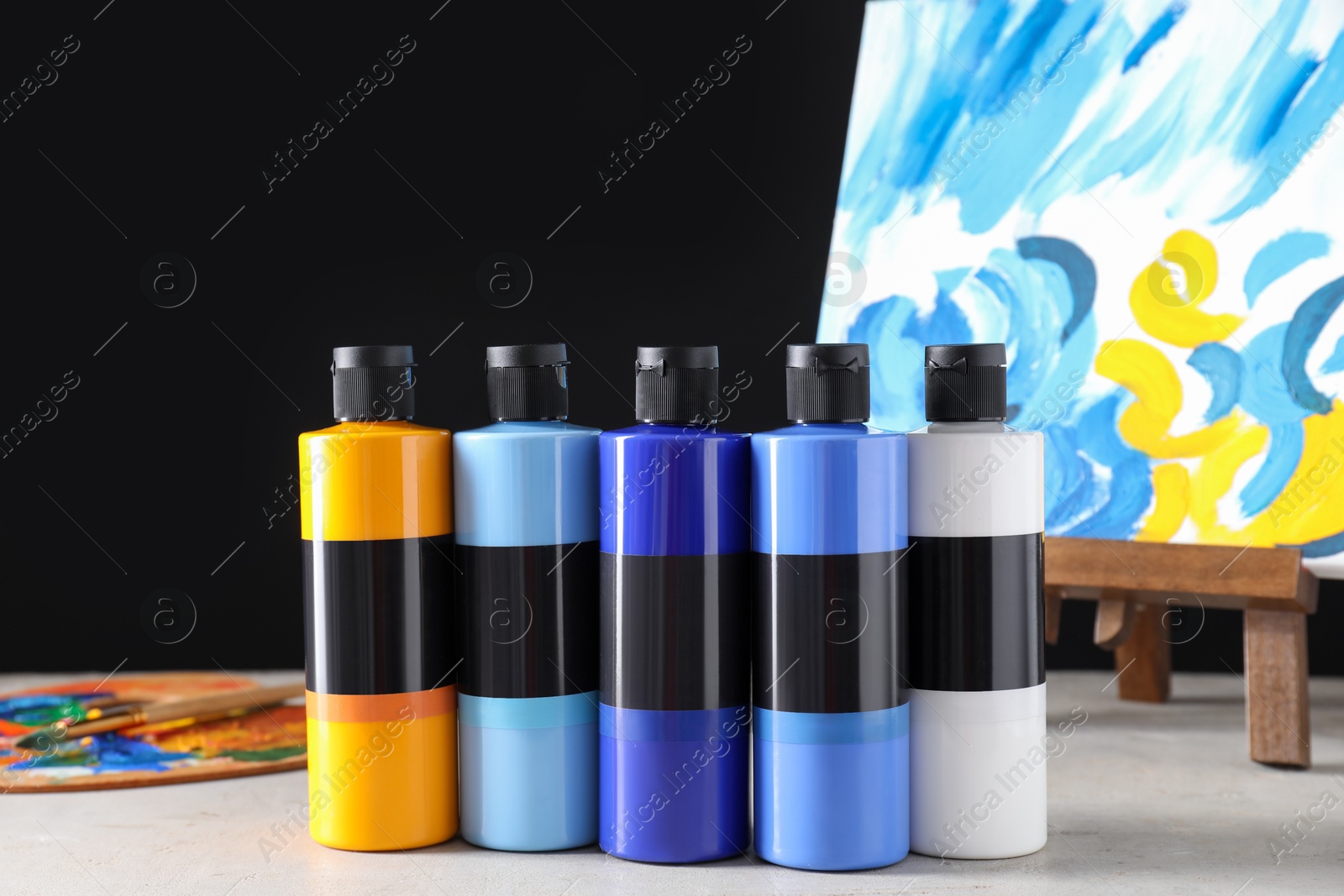 Photo of Acrylic paints of different colors in bottles and small easel with abstract picture on light table