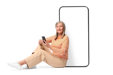 Happy mature woman holding mobile phone and sitting near big smartphone on white background