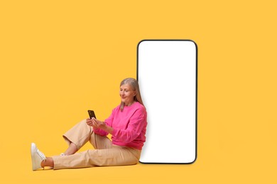 Image of Happy mature woman holding mobile phone and sitting near big smartphone on orange background