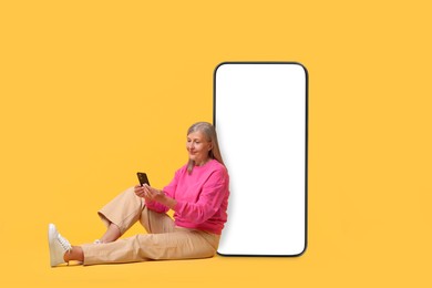 Happy mature woman holding mobile phone and sitting near big smartphone on orange background