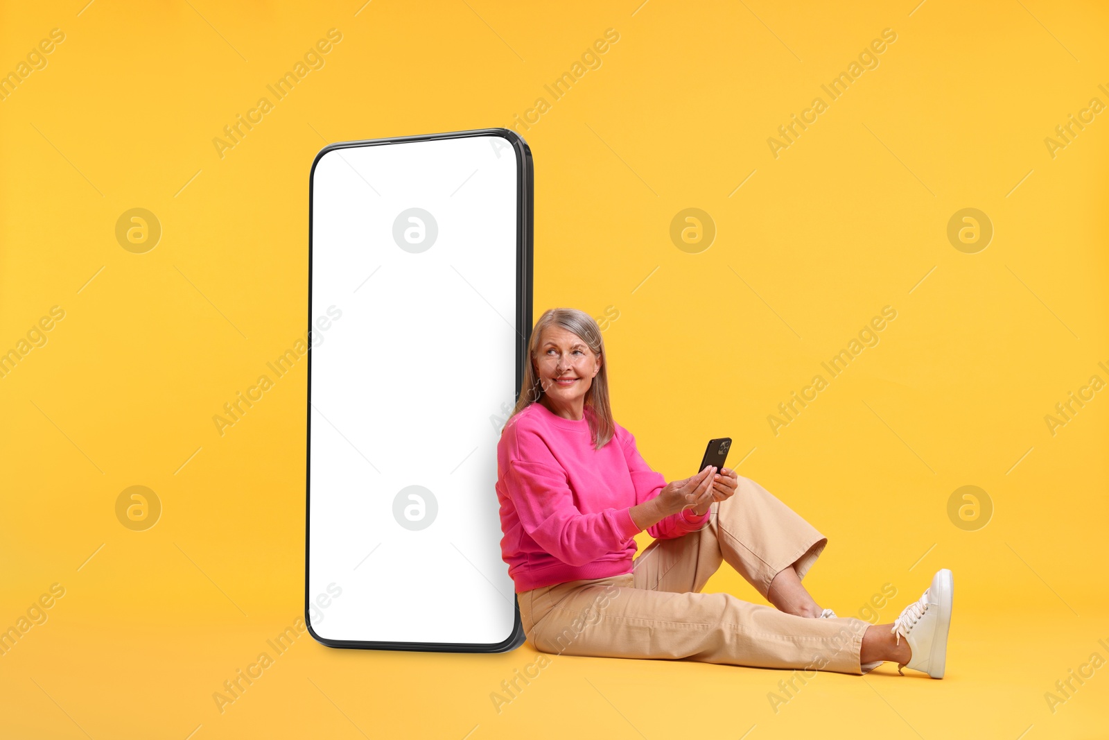 Image of Happy mature woman holding mobile phone and sitting near big smartphone on orange background