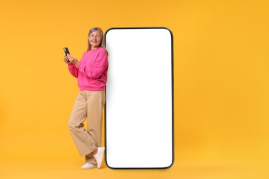 Image of Happy mature woman holding mobile phone near big smartphone on orange background