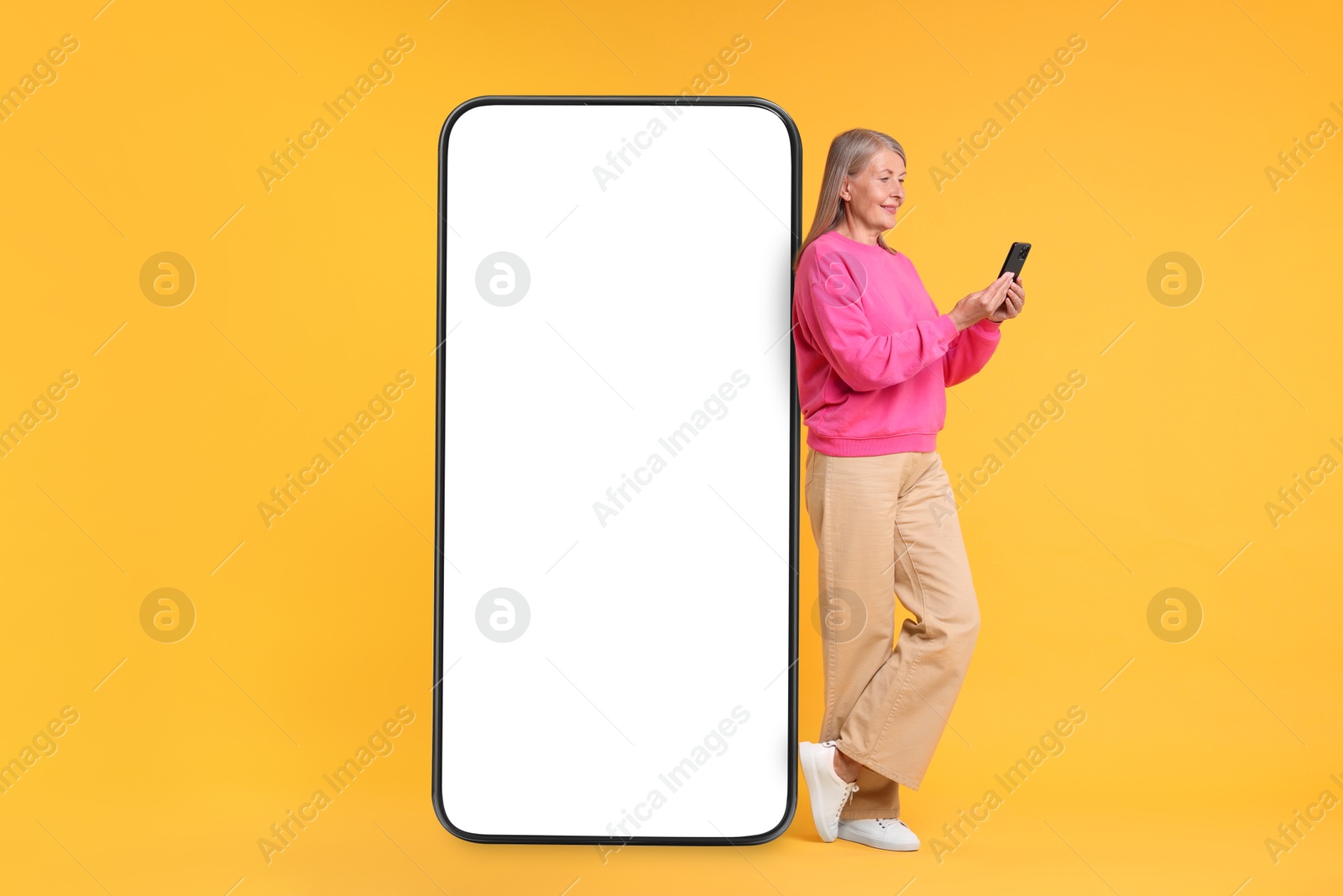 Image of Happy mature woman holding mobile phone near big smartphone on orange background