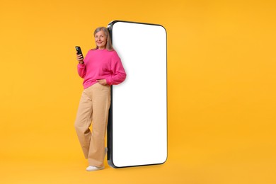 Image of Happy mature woman holding mobile phone near big smartphone on orange background