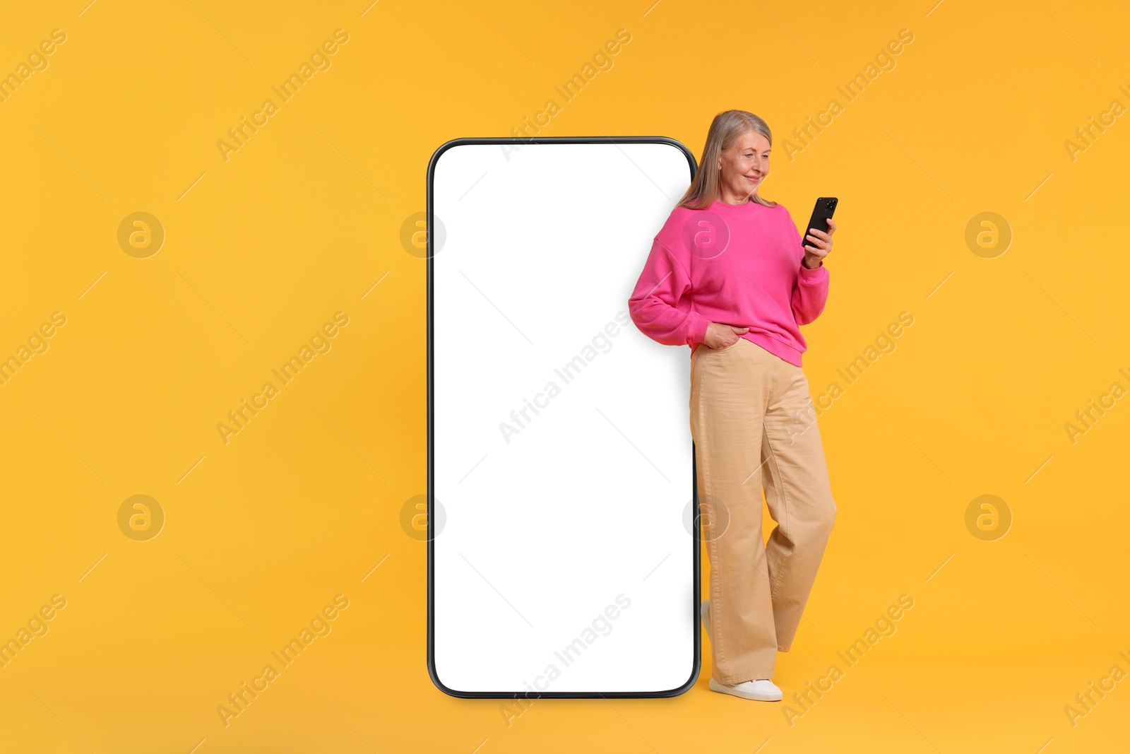 Image of Happy mature woman holding mobile phone near big smartphone on orange background