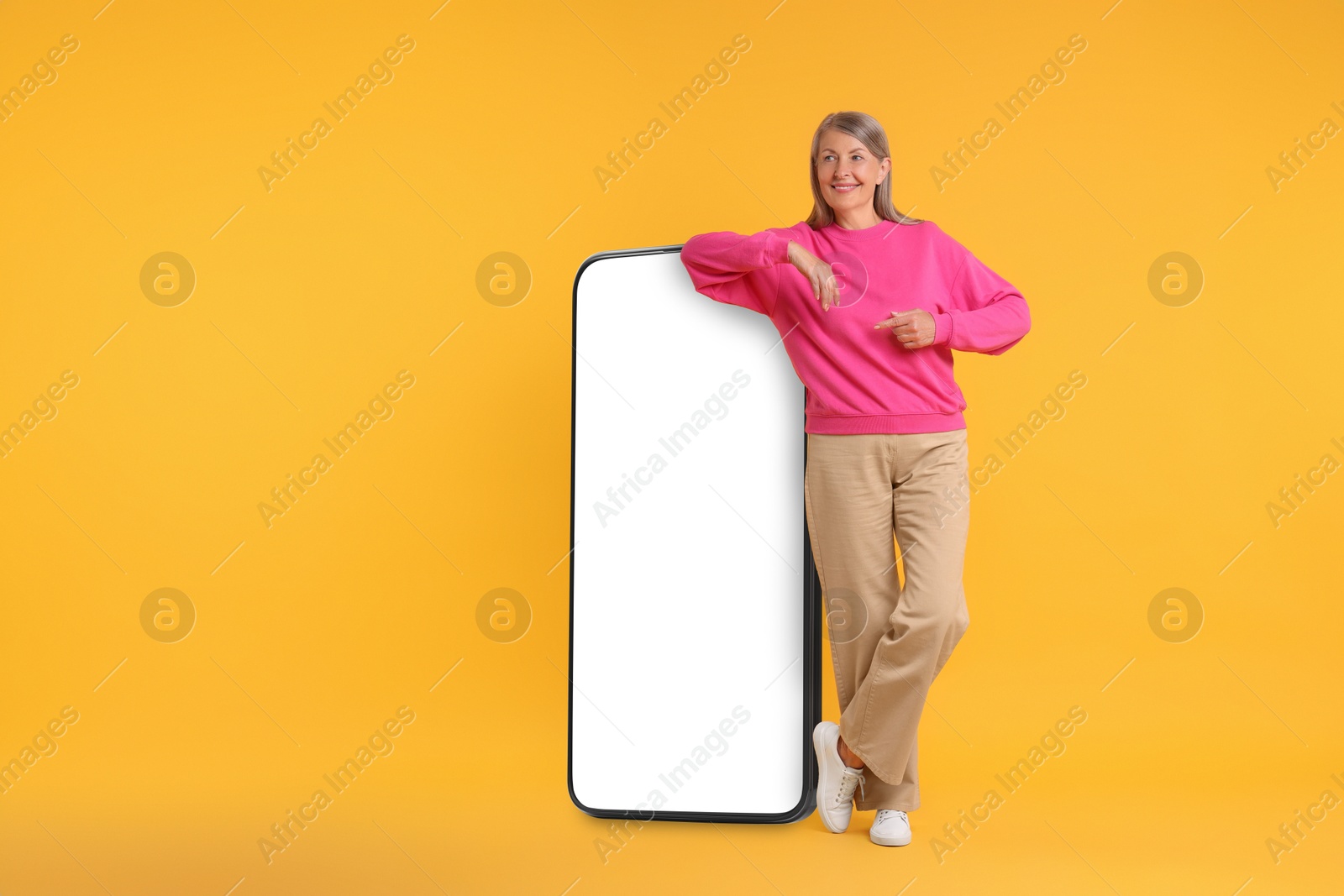 Image of Happy mature woman pointing at big mobile phone on orange background