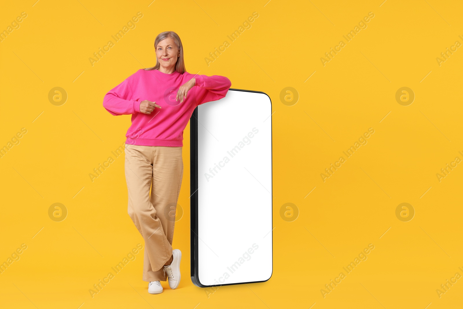 Image of Happy mature woman pointing at big mobile phone on orange background