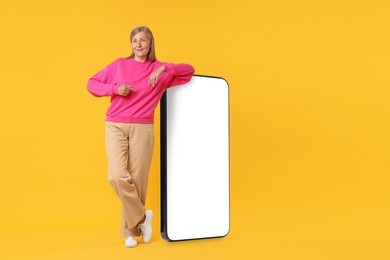 Image of Happy mature woman pointing at big mobile phone on orange background
