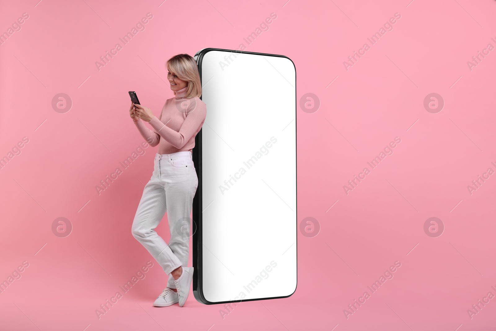 Image of Happy woman holding mobile phone near big smartphone on pink background