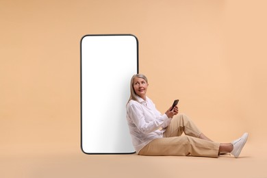 Happy mature woman holding mobile phone and sitting near big smartphone on dark beige background