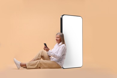 Happy mature woman holding mobile phone and sitting near big smartphone on dark beige background