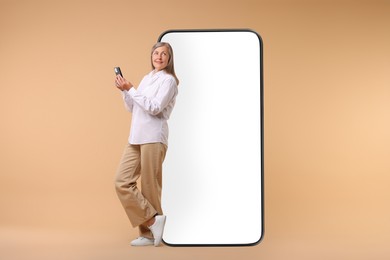 Image of Happy mature woman holding mobile phone near big smartphone on dark beige background