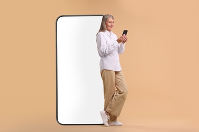 Happy mature woman holding mobile phone near big smartphone on dark beige background