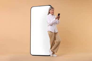 Happy mature woman holding mobile phone near big smartphone on dark beige background