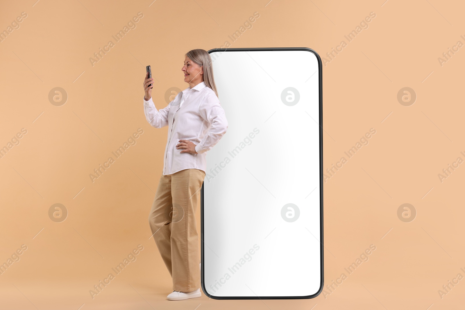 Image of Happy mature woman holding mobile phone near big smartphone on dark beige background