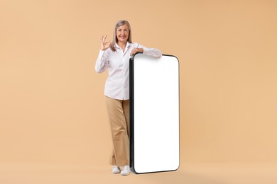 Happy mature woman showing Ok gesture near big mobile phone against dark beige background