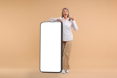 Image of Happy mature woman leaning on big mobile phone against dark beige background