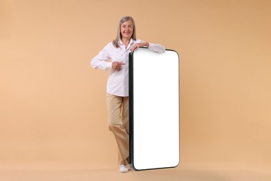 Happy mature woman pointing at big mobile phone on dark beige background