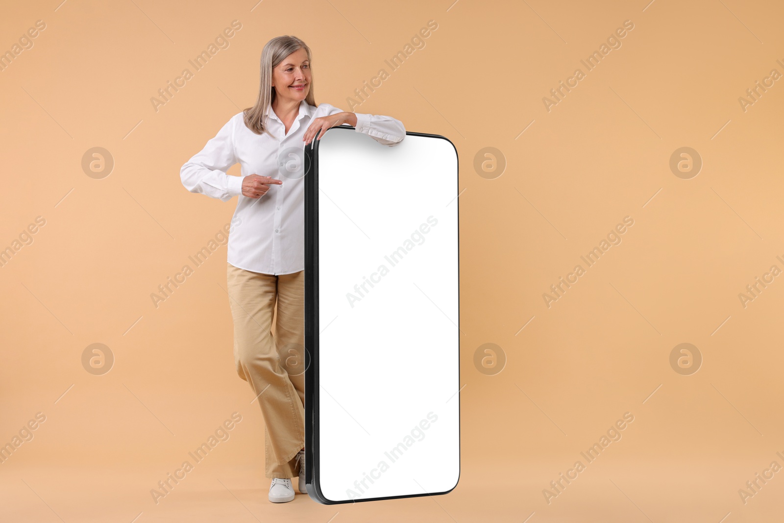 Image of Happy mature woman pointing at big mobile phone on dark beige background