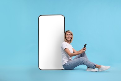 Happy woman holding mobile phone and sitting near big smartphone on light blue background