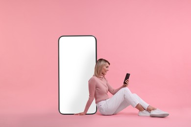Image of Happy woman holding mobile phone and sitting near big smartphone on pink background