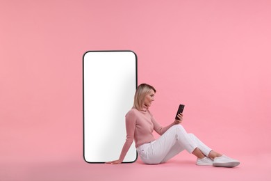 Happy woman holding mobile phone and sitting near big smartphone on pink background