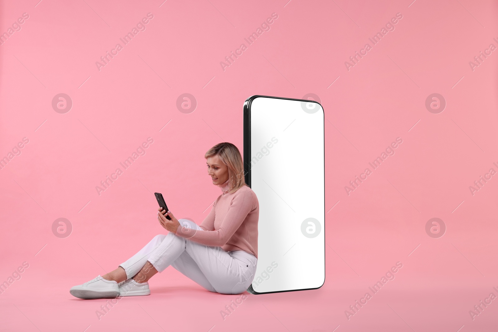 Image of Happy woman holding mobile phone and sitting near big smartphone on pink background