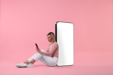 Image of Happy woman holding mobile phone and sitting near big smartphone on pink background