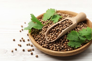 Photo of Dried coriander seeds with green leaves in bowl and scoop on wooden table, closeup