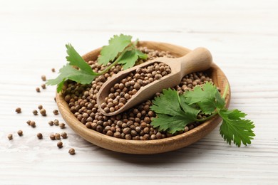 Photo of Dried coriander seeds with green leaves in bowl and scoop on wooden table, closeup