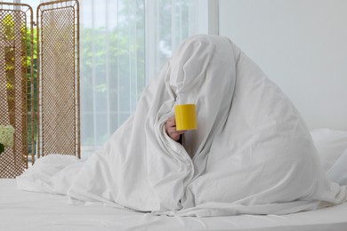 Photo of Bedtime. Woman wrapped in blanket holding cup of drink on bed indoors