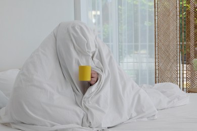 Photo of Bedtime. Woman wrapped in blanket holding cup of drink on bed indoors